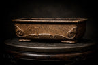 Dark brown rectangular pot with scrolling side relief displayed on a round wooden stand