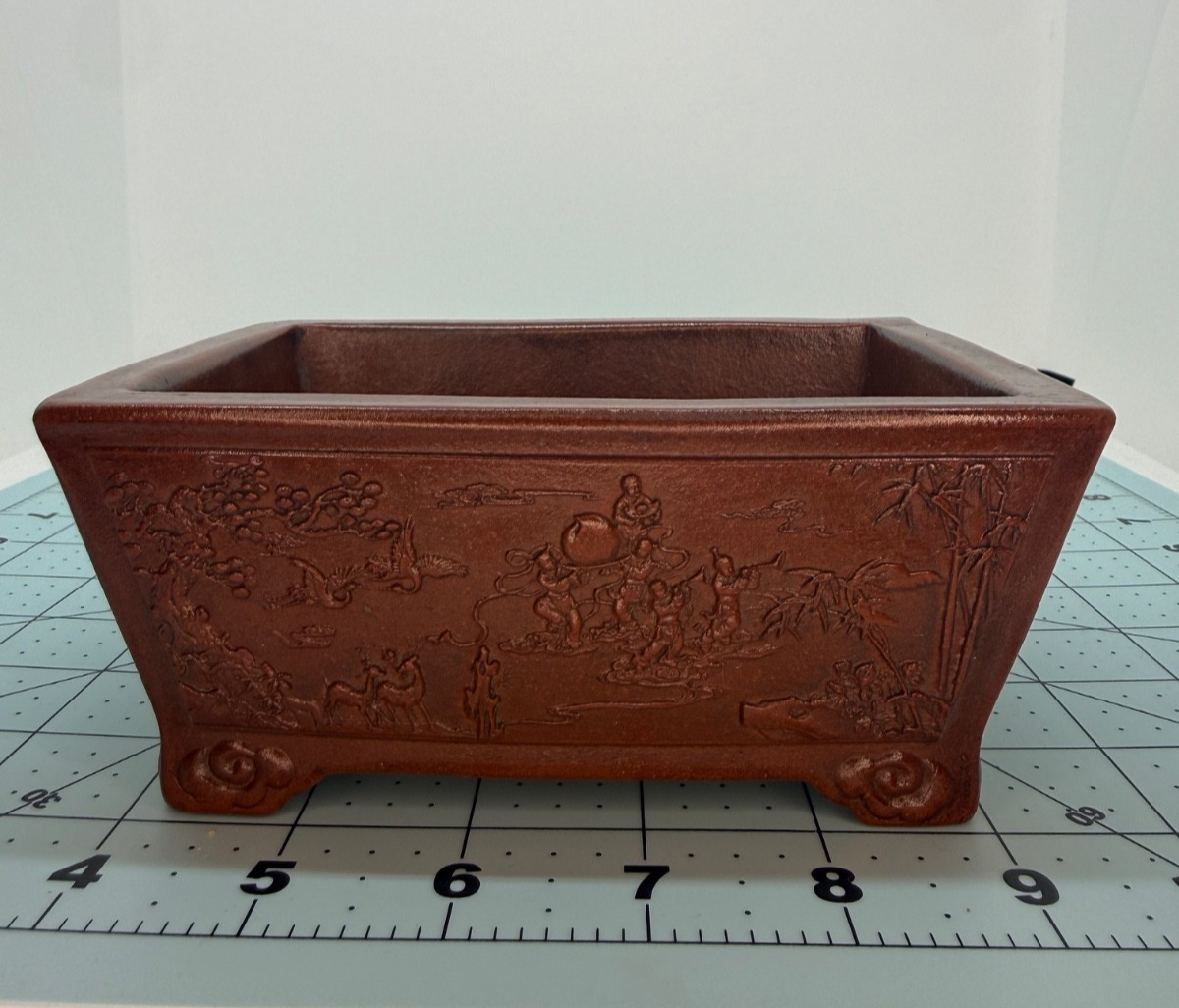 Front view of the reddish-brown pot showing detailed landscape decoration and curled feet