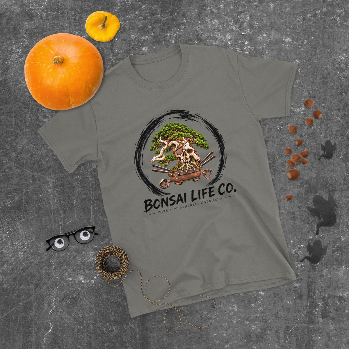 charcoal gray T-shirt with green bonsai and pirate skull graphic inside a black circle with Bonsai Life Co. lettering, gray flat lay with pumpkins