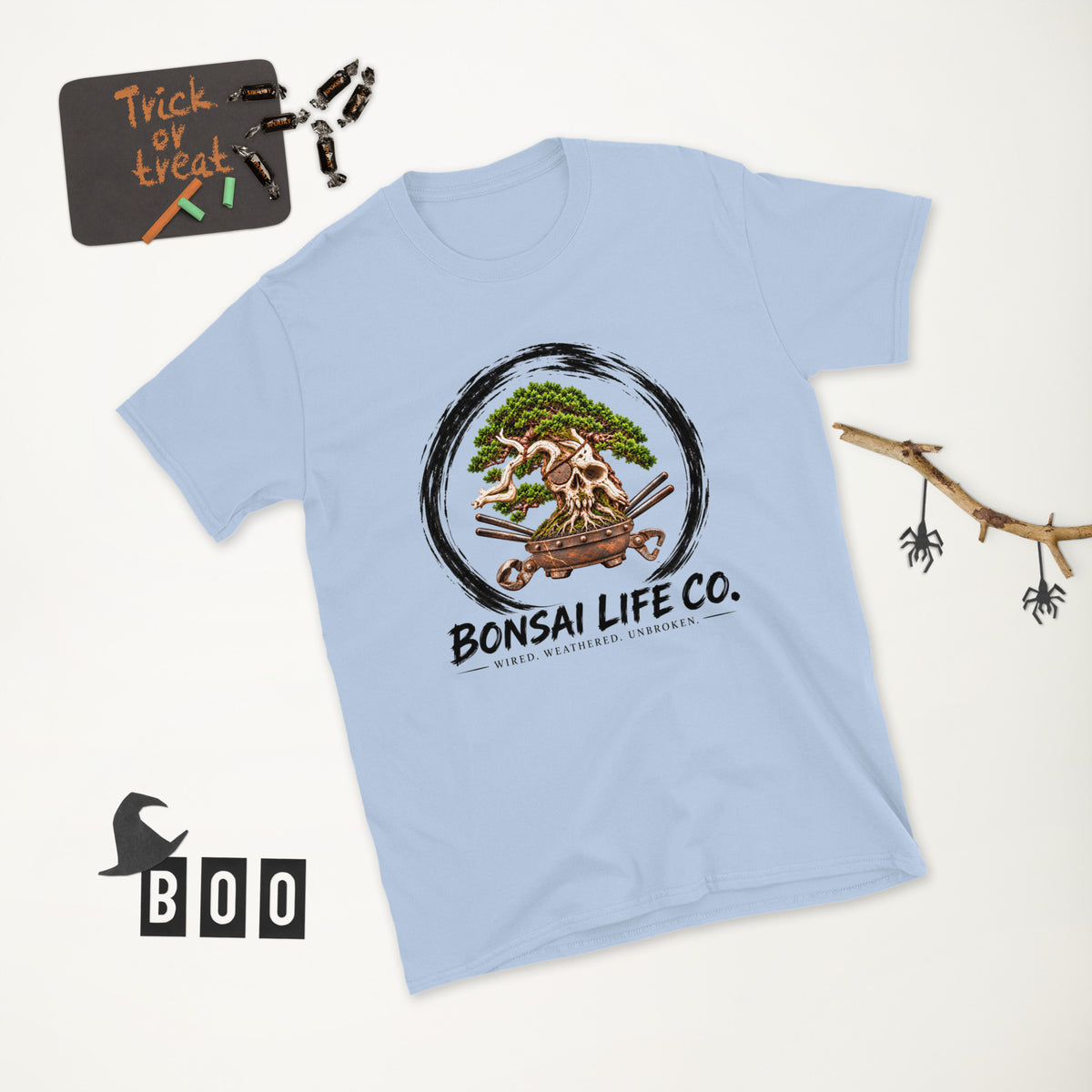 light blue T-shirt with green bonsai and pirate skull graphic inside a black circle with Bonsai Life Co. lettering, white Halloween flat lay