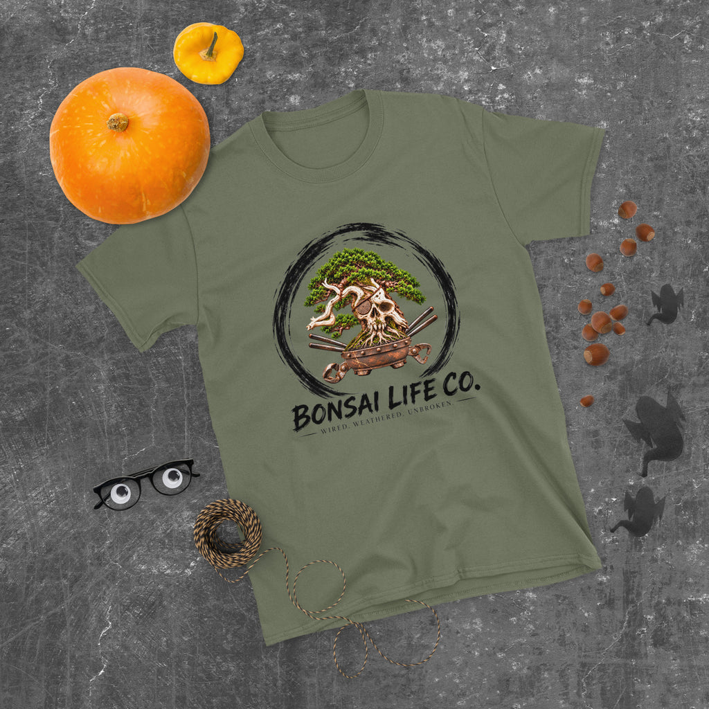 muted olive green T-shirt with green bonsai and pirate skull graphic inside a black circle with Bonsai Life Co. lettering, gray flat lay with pumpkins