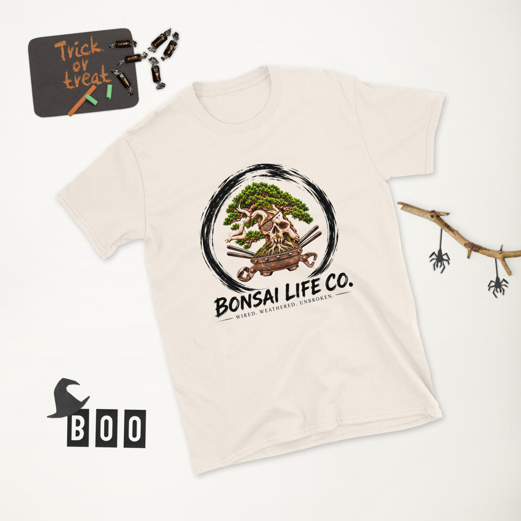 cream T-shirt with green bonsai and pirate skull graphic inside a black circle with Bonsai Life Co. lettering, white Halloween flat lay