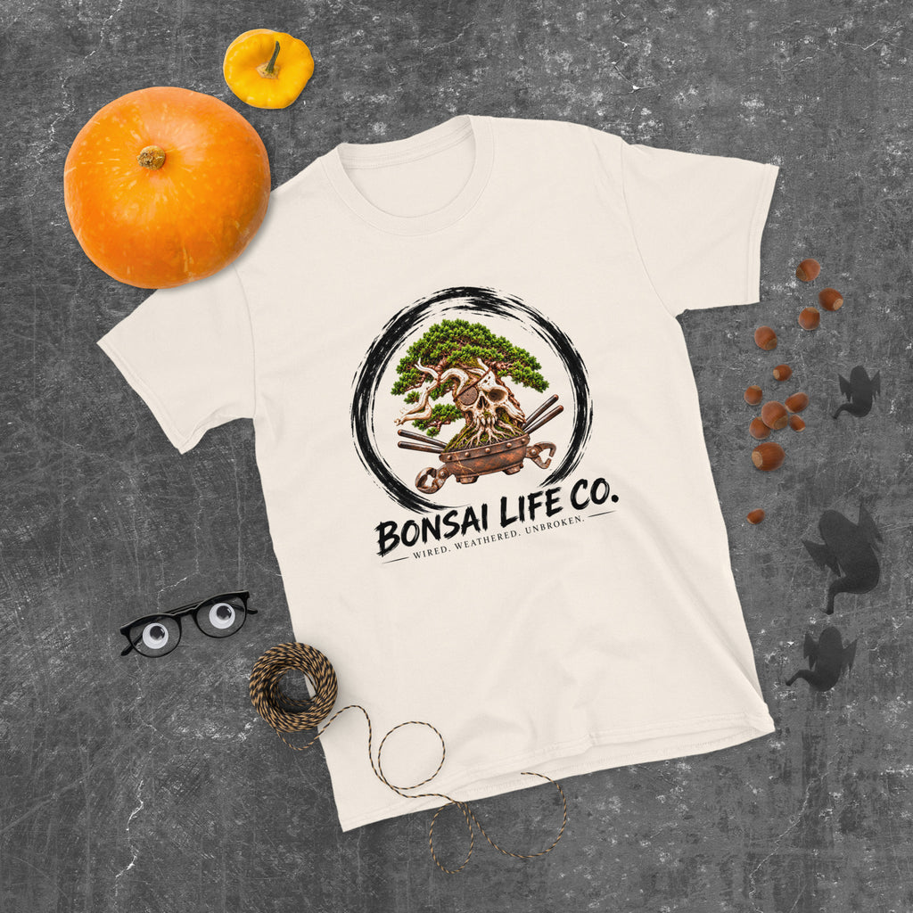 cream T-shirt with green bonsai and pirate skull graphic inside a black circle with Bonsai Life Co. lettering, gray flat lay with pumpkins