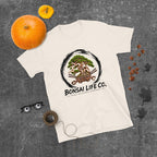 cream T-shirt with green bonsai and pirate skull graphic inside a black circle with Bonsai Life Co. lettering, gray flat lay with pumpkins