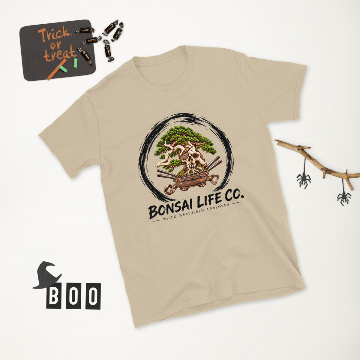 sand beige T-shirt with green bonsai and pirate skull graphic inside a black circle with Bonsai Life Co. lettering, white Halloween flat lay