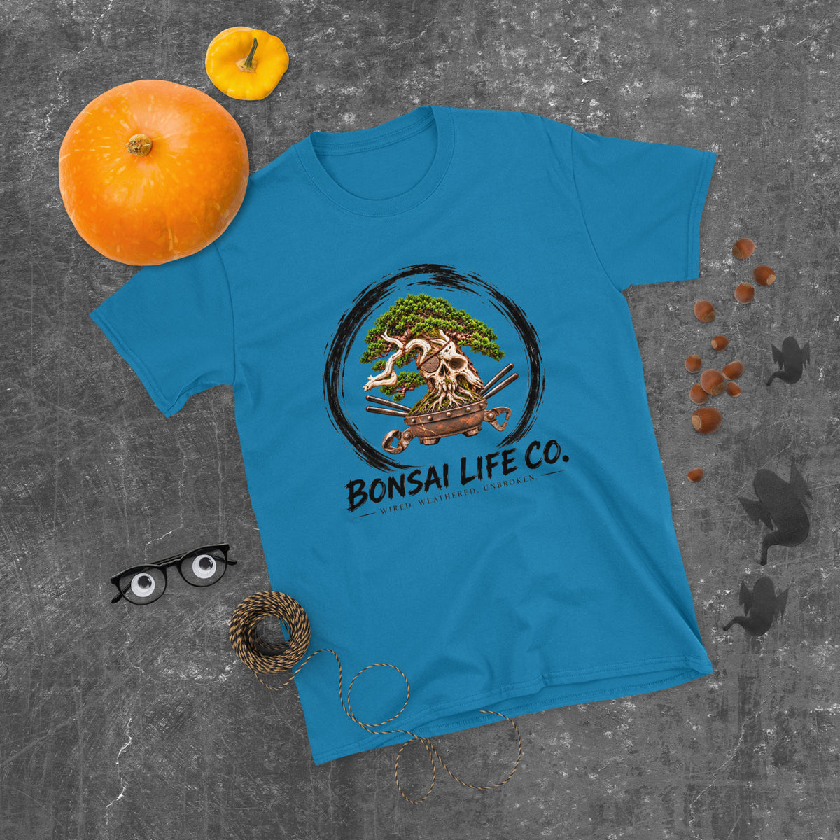 sapphire blue T-shirt with green bonsai and pirate skull graphic inside a black circle with Bonsai Life Co. lettering, gray flat lay with pumpkins