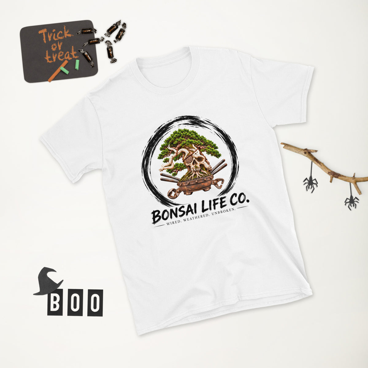 white T-shirt with green bonsai and pirate skull graphic inside a black circle with Bonsai Life Co. lettering, white Halloween flat lay