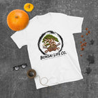 white T-shirt with green bonsai and pirate skull graphic inside a black circle with Bonsai Life Co. lettering, gray flat lay with pumpkins