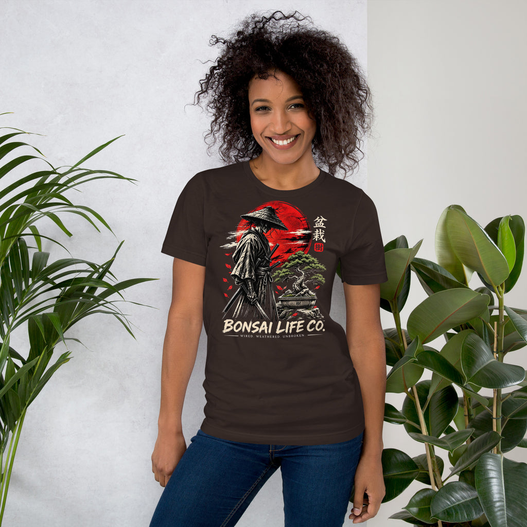 Model wearing dark brown T-shirt with samurai beside a green bonsai and red sun with Bonsai Life Co. lettering, beside leafy plants