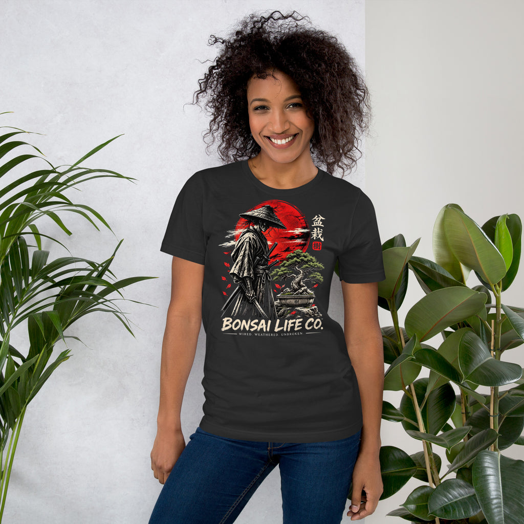 Model wearing dark gray T-shirt with samurai beside a green bonsai and red sun with Bonsai Life Co. lettering, beside leafy plants