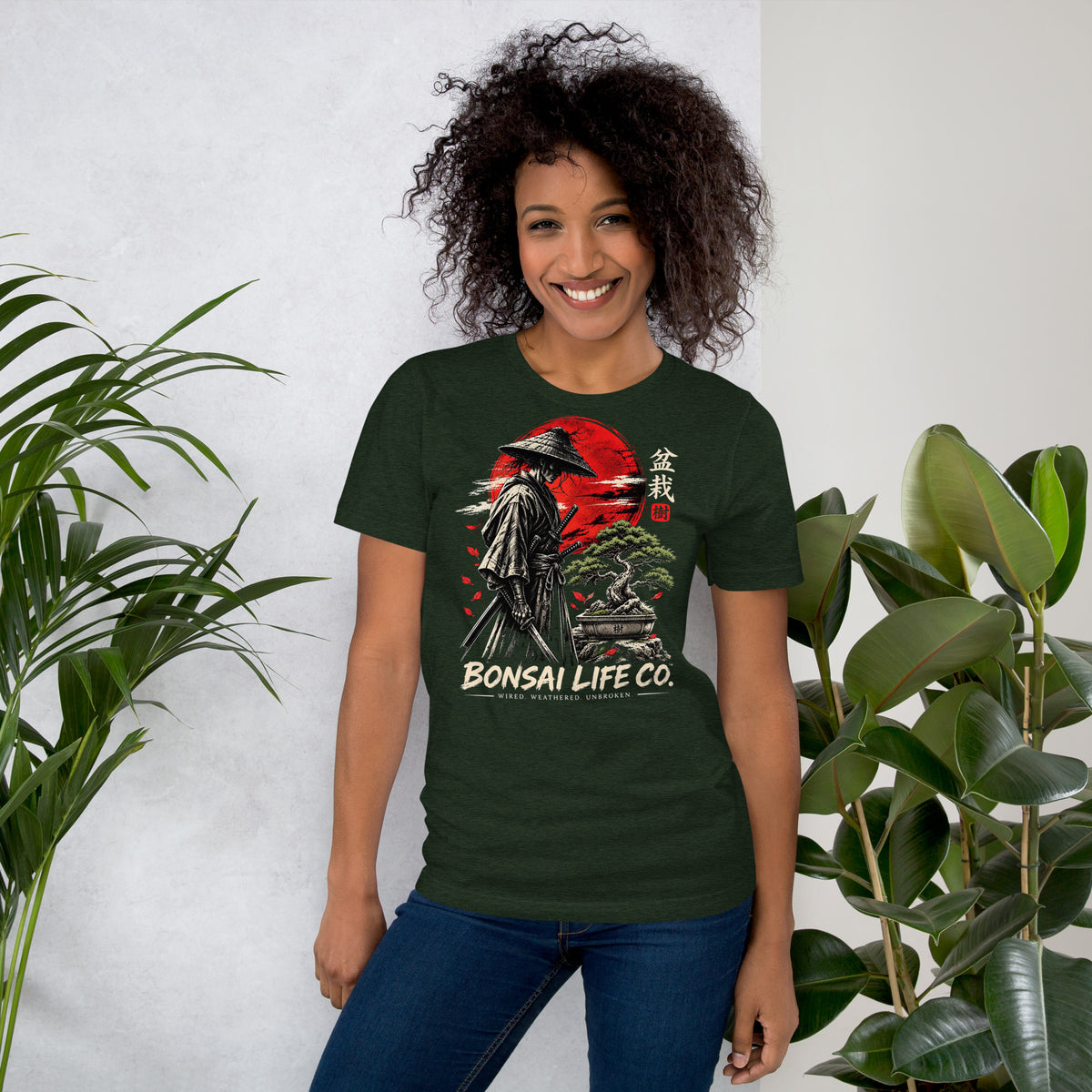 Model wearing heathered dark green T-shirt with samurai beside a green bonsai and red sun with Bonsai Life Co. lettering, beside leafy plants