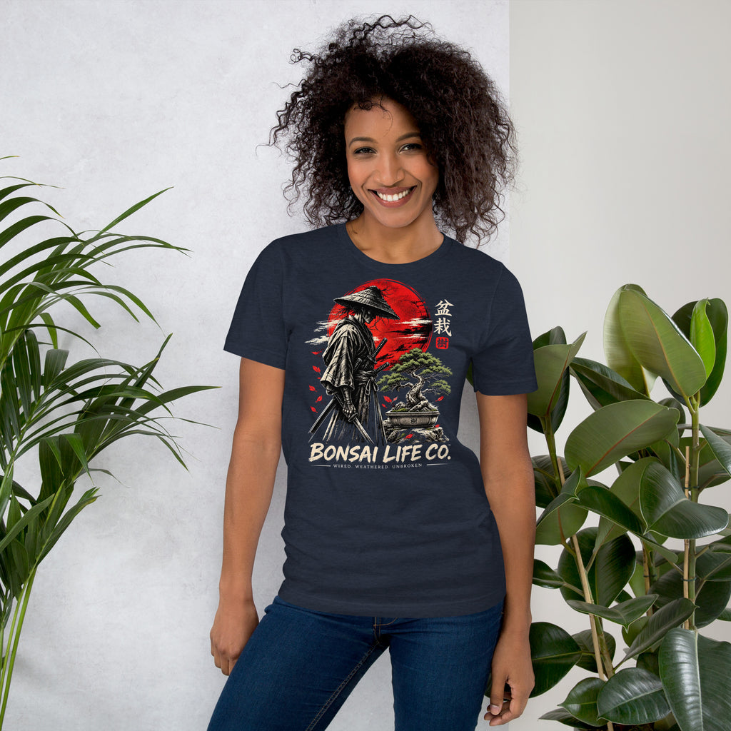 Model wearing heathered navy blue T-shirt with samurai beside a green bonsai and red sun with Bonsai Life Co. lettering, beside leafy plants