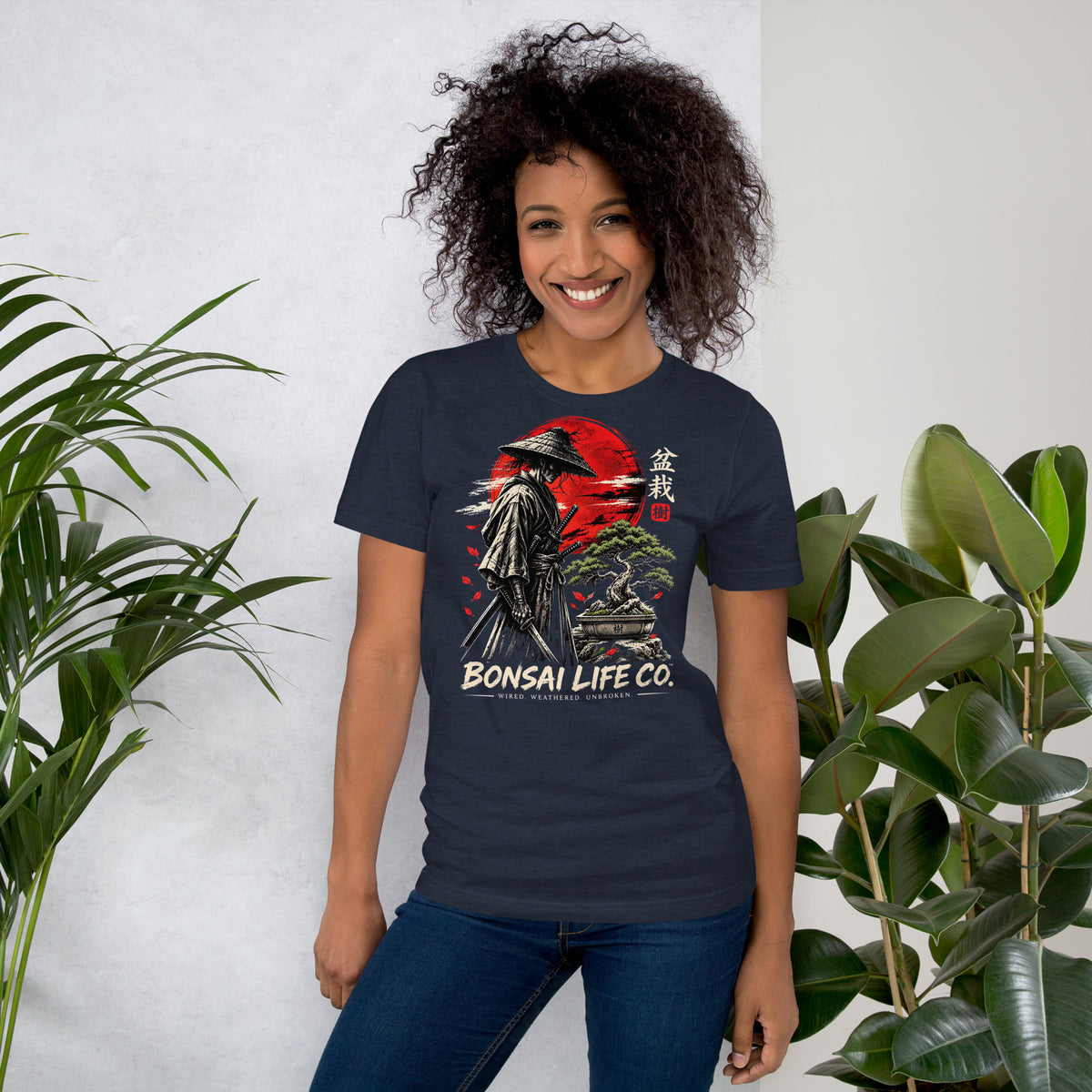 Model wearing heathered navy blue T-shirt with samurai beside a green bonsai and red sun with Bonsai Life Co. lettering, beside leafy plants
