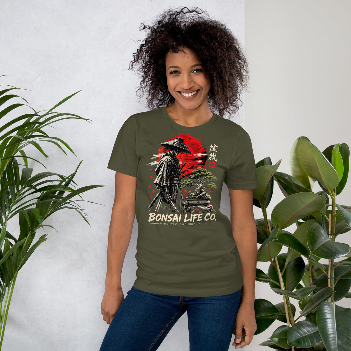 Model wearing muted olive green T-shirt with samurai beside a green bonsai and red sun with Bonsai Life Co. lettering, beside leafy plants