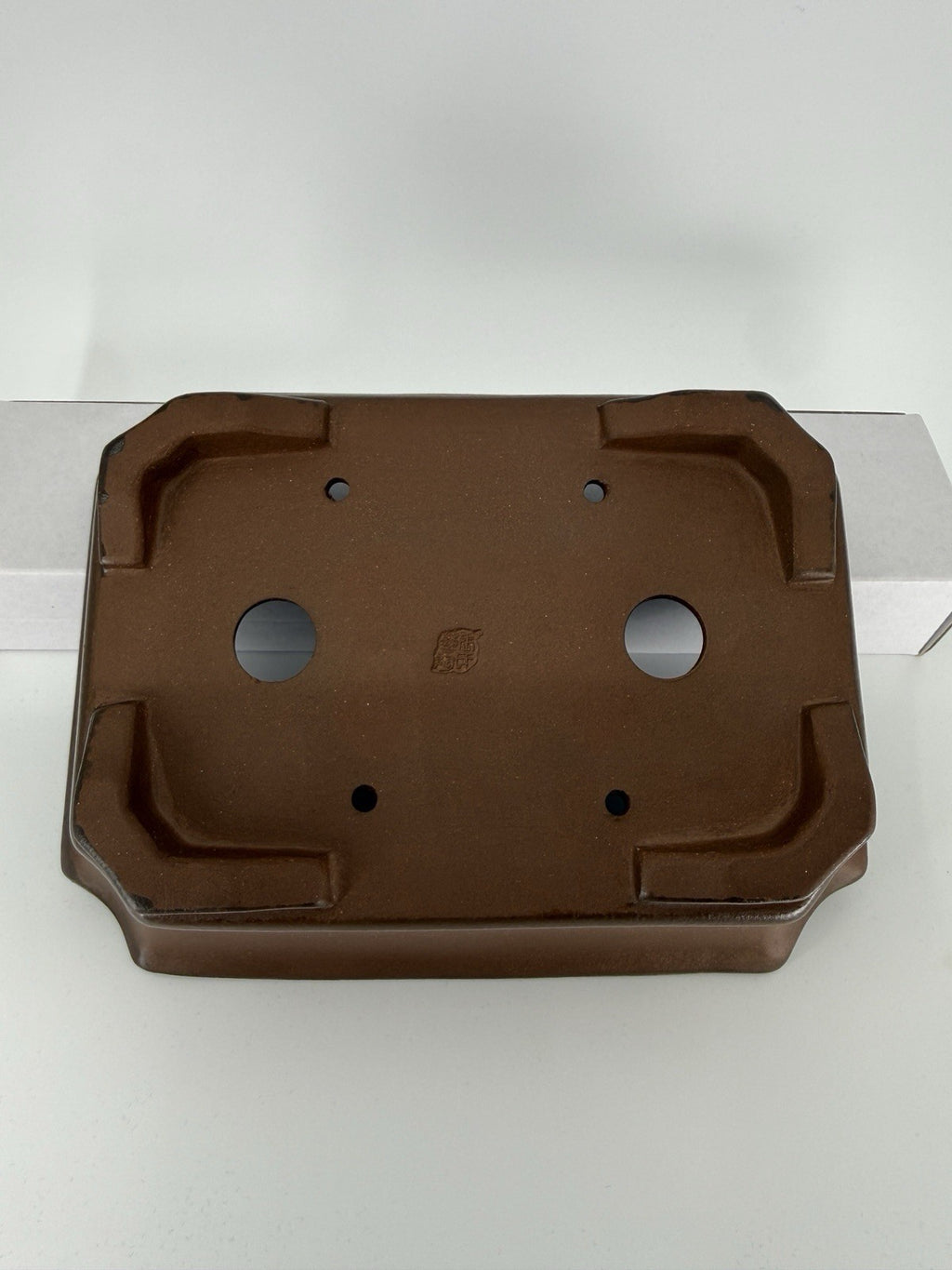 Underside of the brown rectangular pot showing four corner feet, drainage openings and an impressed seal