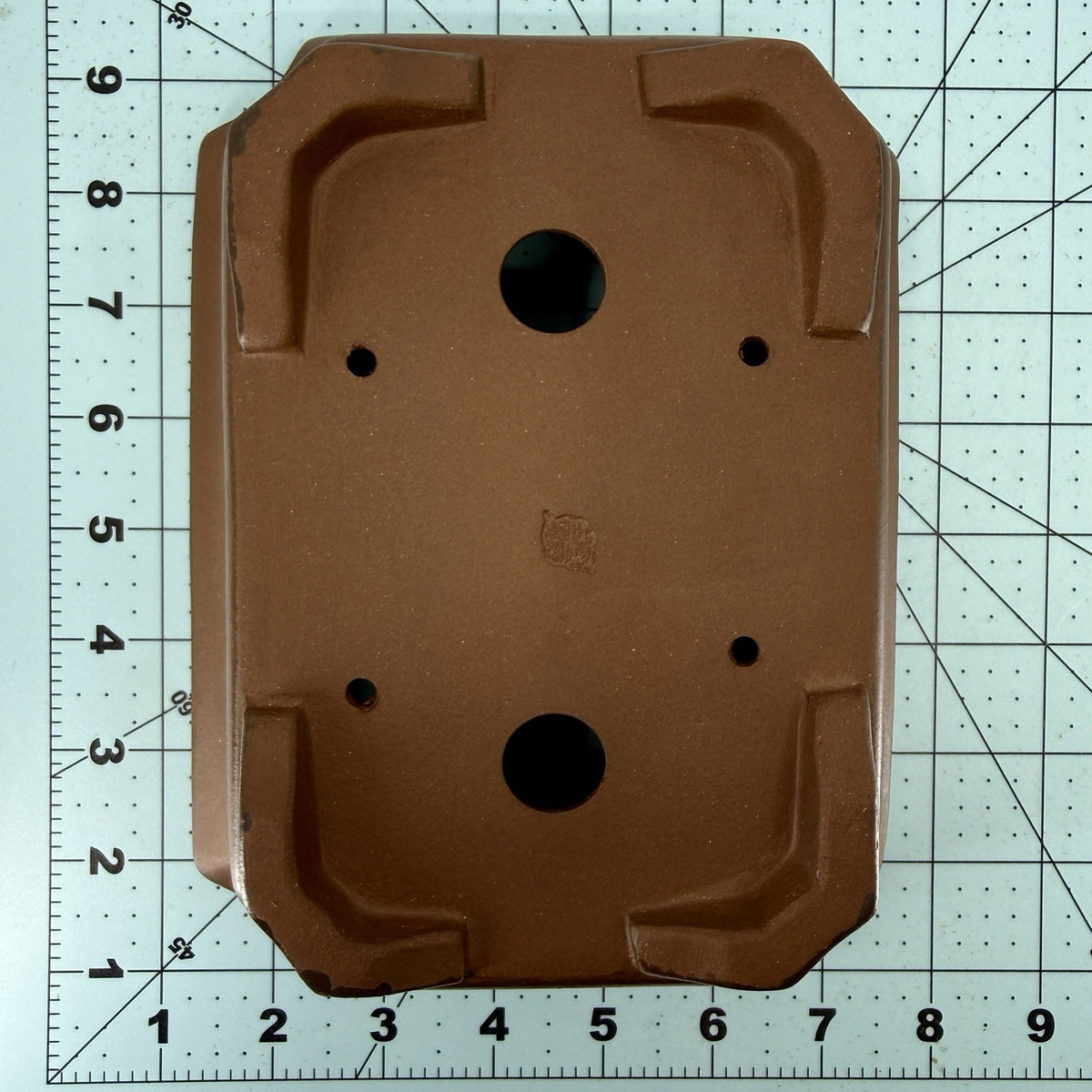 Brown rectangular pot base on a gridded measuring mat, showing drainage holes and corner feet